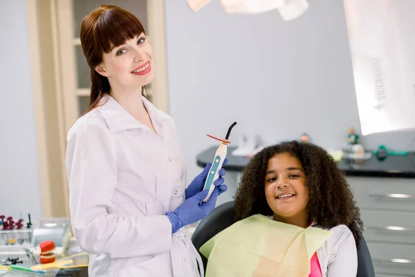 Child at dentists office. Little patient, mixed raced curly girl, sitting in dentistry chair, looking at camera and smiling. Pretty female dentist posing to camera with UV polymerization lamp — Zdjęcie stockowe