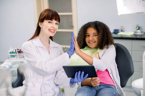 Medicine, pediatric dentistry and oral care concept. Happy female dentist and curly mixed raced kid girl, having fun and giving high five, while holding tablet pc computer — Fotografia de Stock