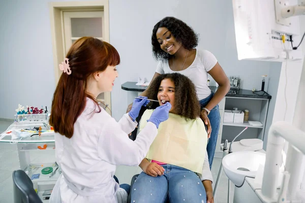 Young smiling African woman mother with her cute preteen daughter, visit their pediatric dentist for regular teeth checkup at stomatology clinic. Child with mom at dentists office — Foto Stock