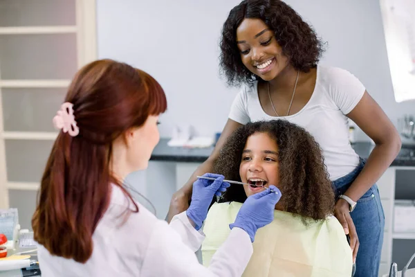 Dental treatment without fear. Female dentist in whilte uniform and gloves, examining teeth of little African American girl, while her mother supporting her behind, at modern pediatric dental clinic — Zdjęcie stockowe