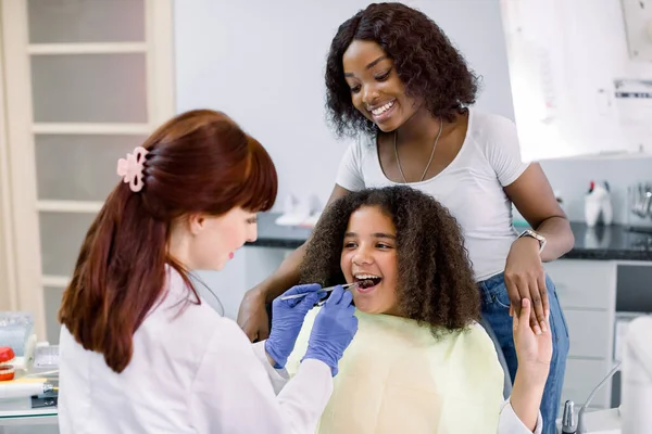 Little mixed raced school girl with curly hair, visiting dentist for checkup or caries treatment, sitting in dental chair and holding hand of her pretty African mom. Female dentist makes examination. — Zdjęcie stockowe