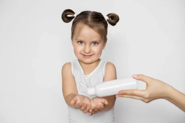 Uma menina de três anos de idade com cabelo loiro, de pé sobre fundo branco isolado, olhando para a câmera com sorriso, enquanto a mão de sua mãe derramando leite creme ou óleo corporal em suas mãos — Fotografia de Stock