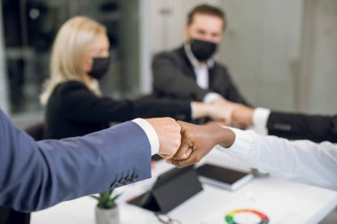 Close up cropped image of happy business coworkers with face protective masks, bumping their fists after successful meeting in the office. Focus on bumping fists of African lady and Caucasian man clipart