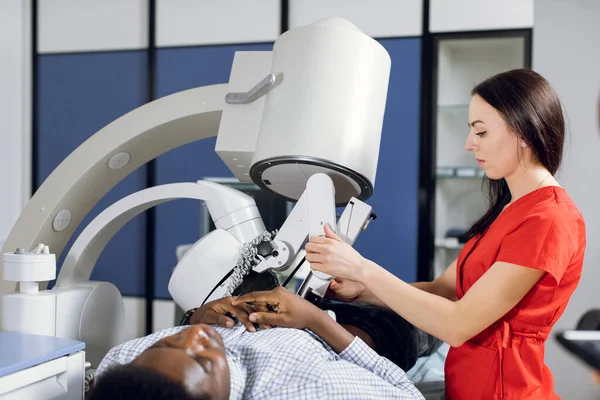 Close up side view of high-skilled female doctor in red uniform, performing lithotripsy, using modern lithotripter to break up kidney stones of male African patient with high-intensity acoustic pulse — Stock Photo, Image