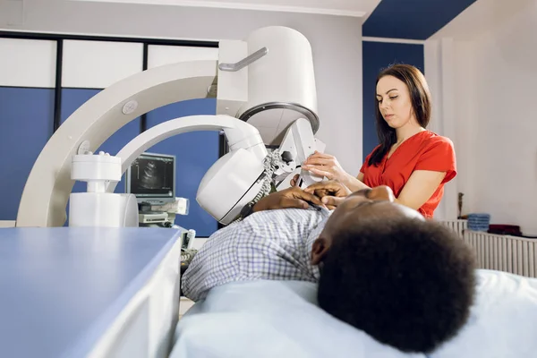 Procedure of extracorporeal shock wave lithotripsy in modern urology medical center. Pretty Caucasian woman doctor providing stone treatment for black male patient using lithotripter — Stock Photo, Image