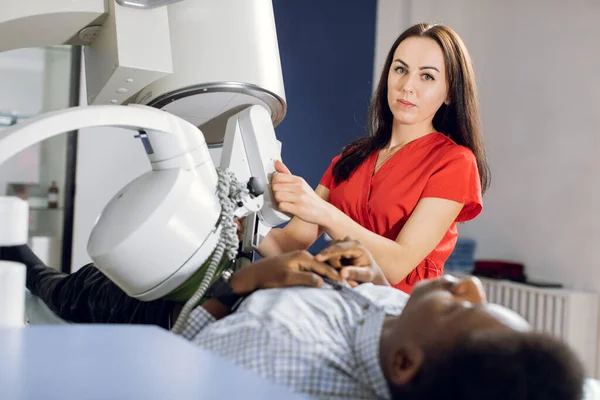 Portrait of young attractive woman doctor, looking at camera, while providing lithotripsy procedure for her male African American patient with modern ultrasonic lithotriptor to break up stones — Stock Photo, Image