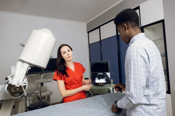 Jovem médico caucasiano feminino na clínica moderna, segurando tablet pc e mostrando ao seu paciente africano masculino ultra-sonografia dos rins e órgãos internos e explicando maneiras de tratamento — Fotografia de Stock