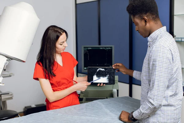 Professional woman ultrasound specialist, holding ipad pc with ultrasound scan image and explaining ways of treatment kidney stones for her young male African American patient — Stock Photo, Image