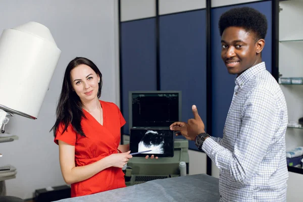 Close up portrait of smiling African man, posing to camera, while visiting his female doctor in modern clinic, holding digital tablet with ultrasound scan. Focus on the woman doctor — Stock Photo, Image