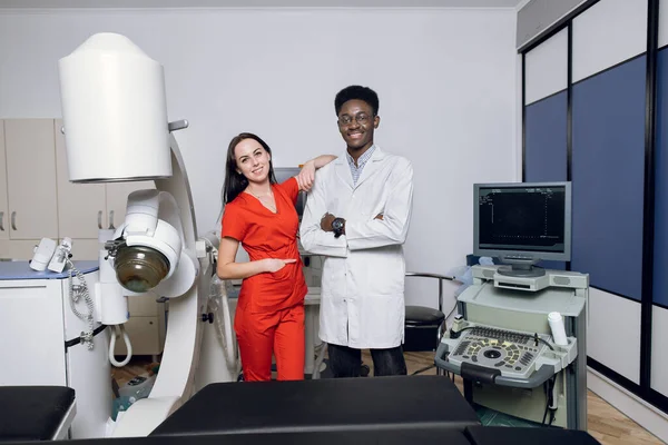 Modern medical center, non-invasive extracorporeal shock wave lithotripsy. Two happy multiethnic male and female doctors, posing to camera near modern lithotripter and ultrasound machine — Stock Photo, Image