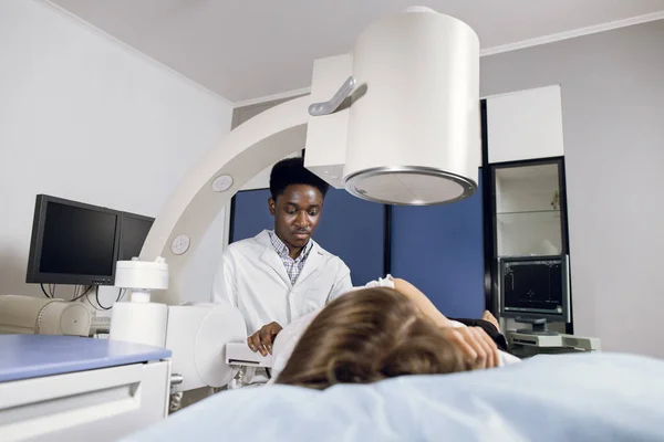 Procedure of extracorporeal shock wave lithotripsy in modern urology medical center. Young African man doctor providing stone treatment or diagnostics for Caucasian female patient using lithotripter — Stock Photo, Image