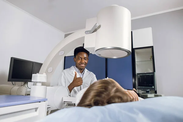 Portrait of young smiling African American man doctor, looking at camera with thumb up, while providing lithotripsy procedure for his female patient with modern ultrasonic lithotriptor — Stock Photo, Image