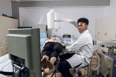 Extracorporeal şok dalga terapisi. Böbrek taşı tedavisi. Afrikalı erkek doktor, kadın hastanın taş pozisyonunu belirlemek için lithotriptorun dijital ekranına bakıyor.