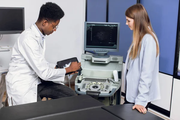 Young male african doctor in modern clinic, holding tablet pc and showing to his female caucasian patient ultrasound scan of kidneys and internal organs and explaining ways of treatment. — Stock Photo, Image