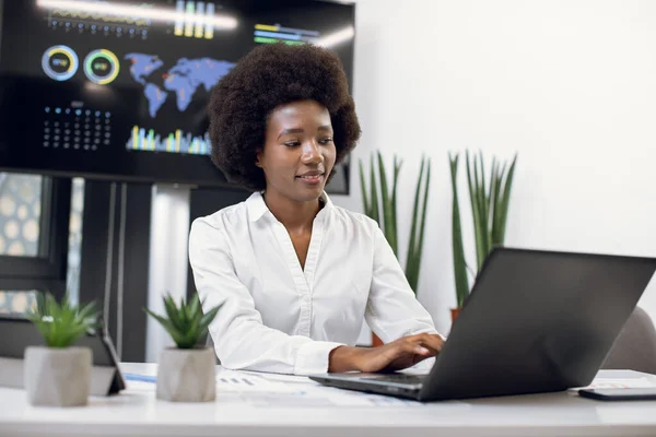 Vue de face de belle femme d'affaires africaine concentrée souriante portant une chemise formelle blanche, avec une coiffure afro, qui tape sur un ordinateur portable dans un bureau moderne. Écran mural numérique derrière — Photo