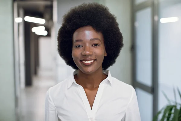 Close-up gezicht portret van mooie glimlachende zwarte jonge vrouw met afrohaar, het dragen van een wit shirt, kijken naar de camera, terwijl ze in een modern kantoorgebouw. Zakenvrouw indoor portret — Stockfoto