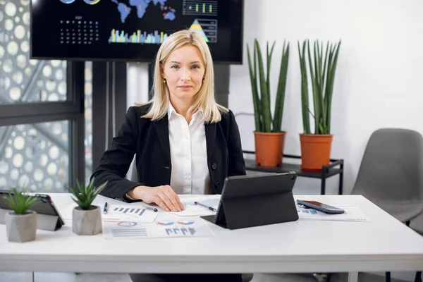 Mujer rubia bastante madura de 40 años en ropa de negocios, director financiero, jefe, sentado a la mesa en la moderna sala de oficina, posando ante la cámara mientras trabaja con la tableta, papeles del proyecto y gráficos —  Fotos de Stock