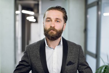 Kendine güvenen genç ofis çalışanı yönetici, resmi kıyafetler giyiyor, kameraya gülümsüyor, modern ışık ofis koridorunun arka planında duruyor. Yüz portresini kapat