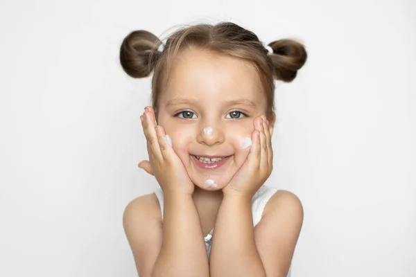 Beyaz bluz giyen beyaz elbiseli beyaz bir bebek, yüzüne nemlendirici krem ya da bebek losyonu sürüyor. Bebek hijyeni ve cilt bakımı kavramı