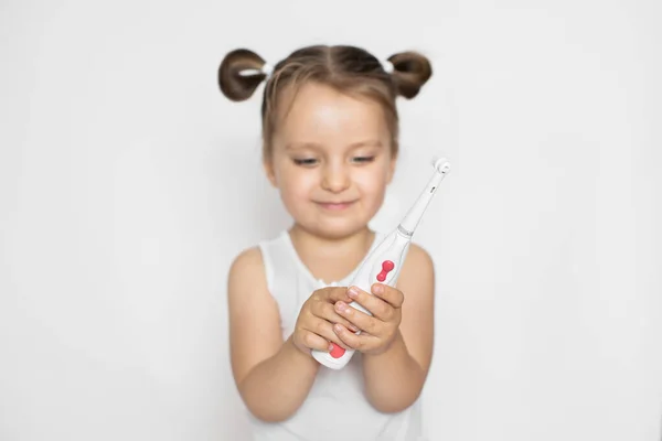 Menina bonito com o cabelo em rabo de cavalo, de pé sobre fundo branco isolado, animado para escovar os dentes com uma escova de dentes elétrica. Feche, copie o espaço. Concentre-se na escova de dentes nas mãos — Fotografia de Stock