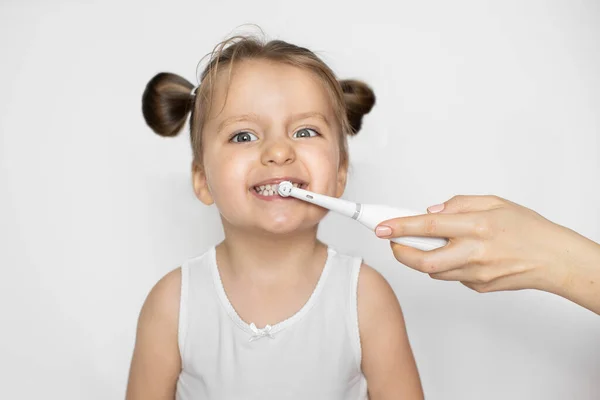 Close up de menina bonito em top branco, feliz por ter os dentes limpos e saudáveis, enquanto a mão de sua mãe limpar os dentes com escova de dentes elétrica. Isolado sobre fundo branco — Fotografia de Stock