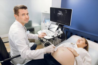 Gülümseyen bir kadın doğum uzmanı ultrasonik taramayla hamile karnı muayene ediyor. Modern annelik merkezindeki doktorunu ziyaret eden mutlu hamile kadın ultrason taraması yapıyor.