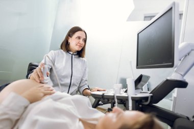 Genç, gülümseyen, çekici kadın doktor klinikte hamile bir kadının karnına 3 boyutlu ultrason yapıyor. Hamile bir kadın için ultrason