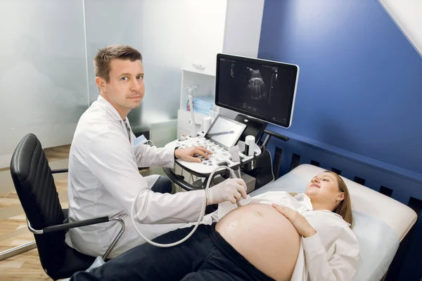 Imagem horizontal de belo jovem médico do sexo masculino, olhando para a câmera, durante a realização de procedimento de diagnóstico de ultra-som em mulheres grávidas na clínica moderna. Conceito de ultrassom, obstetrícia — Fotografia de Stock