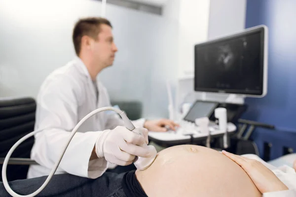 Imagem de perto do médico do sexo masculino que executa o procedimento de ultrassonografia à mulher grávida irreconhecível. Concentre-se no transdutor de ultra-som na barriga da futura mãe — Fotografia de Stock