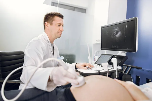 Visão lateral retrato de profissional focado agradável médico masculino caucasiano, fazendo ultra-som para a mulher grávida no hospital moderno. Foco no rosto dos médicos — Fotografia de Stock