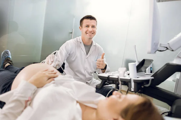 Médico masculino profissional de triagem de mulher grávida por ultra-som, sorrindo e mostrando o polegar para cima. Jovem obstetra examinando barriga grávida por ultra-som na clínica moderna — Fotografia de Stock
