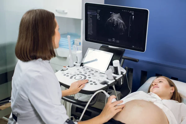 Glückliche Schwangerschaftszeit, Ultraschallkonzept. Hübsche Schwangere beim Ultraschallbild in der pränatalen Klinik, besorgt um die Gesundheit ihres zukünftigen Babys. Ärztin blickt auf den Bildschirm — Stockfoto