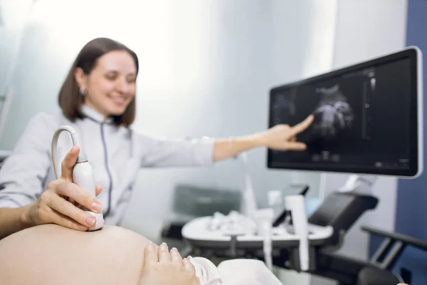 Tiro cortado de uma mulher grávida durante a ecografia na clínica de fertilidade. Médica apontando para a tela da máquina de ultra-som. Concentre-se no transdutor de ultra-som na barriga grávida — Fotografia de Stock