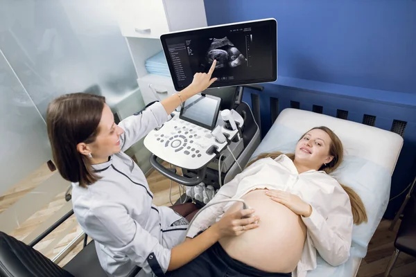 Médico mulher muito sorridente fazendo um diagnóstico de ultra-som para sua jovem paciente grávida feliz, e apontando para a tela, mostrando a imagem ultra-sônica babys. Visão de ângulo superior — Fotografia de Stock