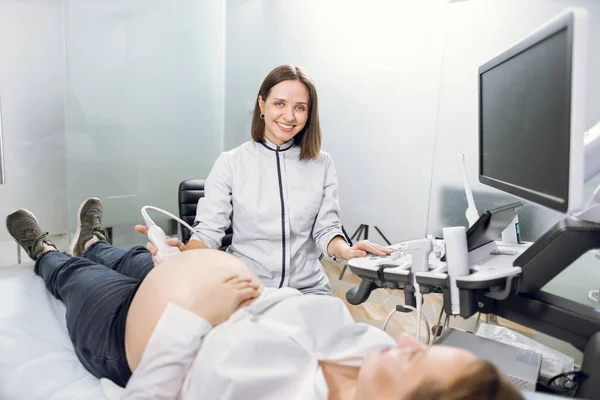 Retrato de una joven y atractiva doctora, posando ante la cámara con sonrisa mientras realizaba un procedimiento de ecografía para su paciente, una joven embarazada. Concepto de exploración por ultrasonido —  Fotos de Stock