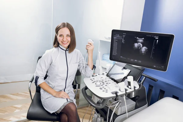 Jovem profissional médico sonógrafo, ginecologista obstetra, sentado nas proximidades moderna máquina de ultra-som scanner e sorrindo para a câmera — Fotografia de Stock