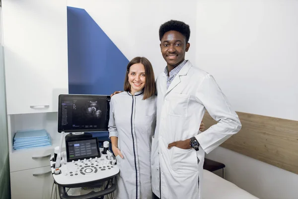 Zwei glücklich lächelnde Ärzte, eine kaukasische Frau und ein afrikanischer Mann, posieren zusammen in der Nähe eines Ultraschallgeräts in einem modernen medizinischen Zentrum und umarmen sich. Ultraschalldiagnostisches Konzept — Stockfoto