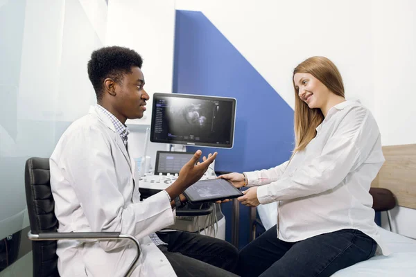 Médico e paciente na sala de ultra-som. Médico masculino africano conversando com sua jovem gestante após o procedimento ultrassônico, contando sobre os resultados do teste de ultra-som mostrando a varredura no tablet — Fotografia de Stock