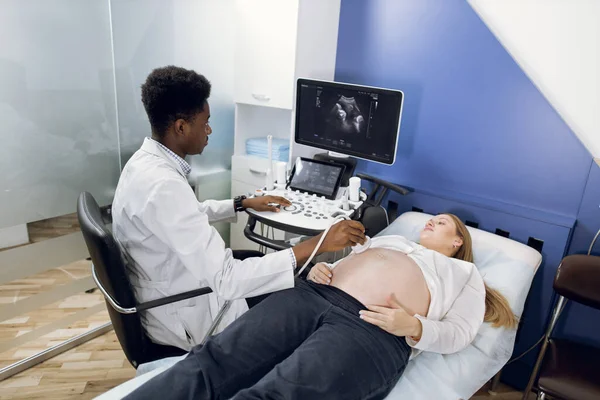 Ultrassom da gravidez. Jovem focado médico masculino Africano altamente qualificado, realizando exame da barriga grávida com dispositivo de ultrassonografia para sua jovem paciente senhora caucasiana — Fotografia de Stock