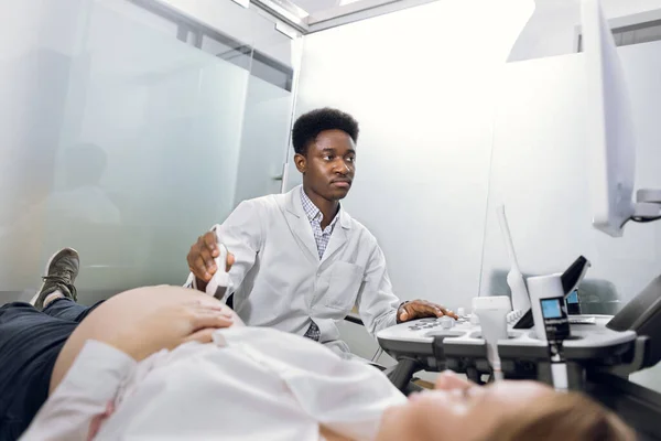 Jovem de pele preta masculina altamente qualificada médica, operando scanner de ultra-som enquanto examina a barriga grávida de sua paciente feminina. Digitalização por ultrassom — Fotografia de Stock