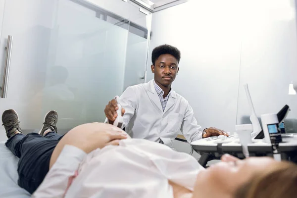 Retrato de jovem profissional do sexo masculino afro-americano médico obstetra, usando scanner de ultra-som e barriga de triagem de mulher grávida. — Fotografia de Stock