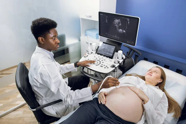 Jovem mulher grávida bonita recebendo ultra-som do médico. Profissional confiante homem Africano médico obstetra, verificando o bebê na barriga com scanner de transdutor de ultra-som. — Fotografia de Stock