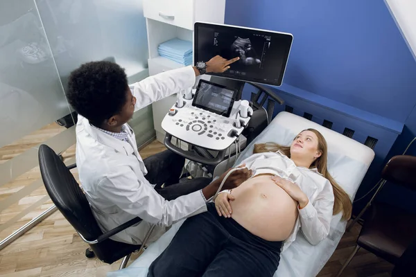Conceito de ultrassom. Visão de ângulo superior da jovem mulher grávida caucasiana deitada no sofá, enquanto um médico afro-americano altamente qualificado faz a varredura de ultasound e mostra o bebê na tela — Fotografia de Stock