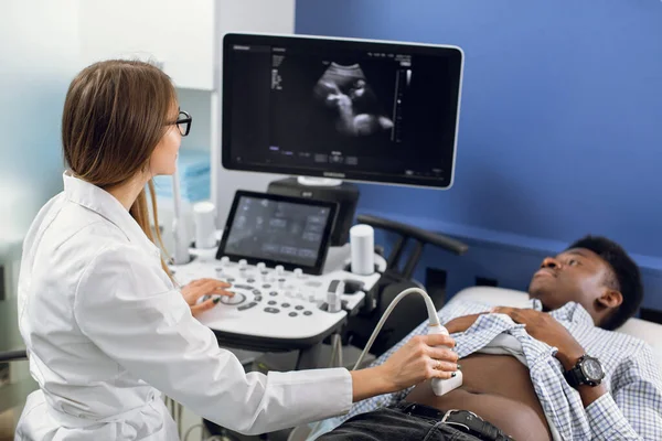 Young Caucasian woman doctor moving ultrasound transducer on African mans belly in hospital. Female doctor making ultra sound of internal organs of her patient.