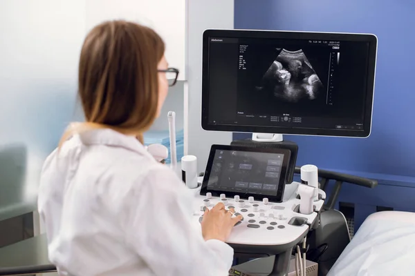 Rückansicht einer jungen kaukasischen Ärztin, die an modernen Ultraschallgeräten arbeitet. Bediener des Ultraschallgeräts sitzt auf dem Bildschirm und bereitet sich auf die Arbeit vor — Stockfoto
