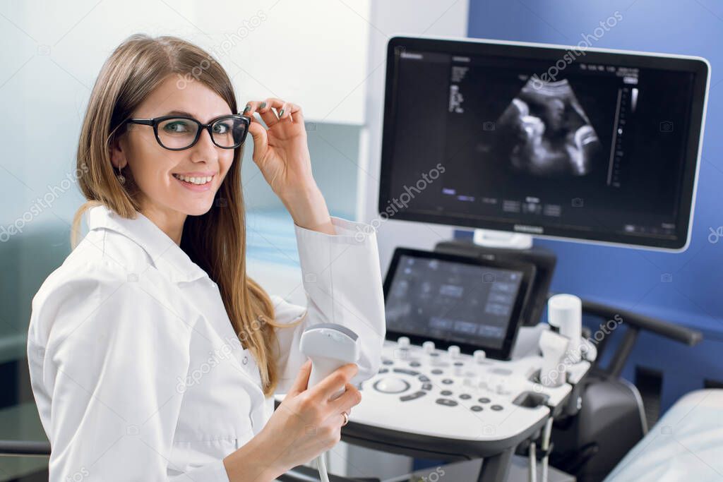 Close up of young happy female doctor in white coat posing to camera with smile while sitting near ultra ultrasound scanning machine in modern light clinic. Ultrasound equipment concept
