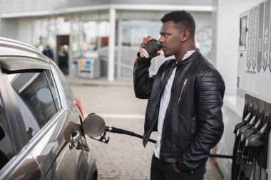 Benzin istasyonunda dikilen, kahve içen, lüks arabalarının deposunu benzin pompasıyla dolduran genç Afrikalı adamın yan görüntüsü.