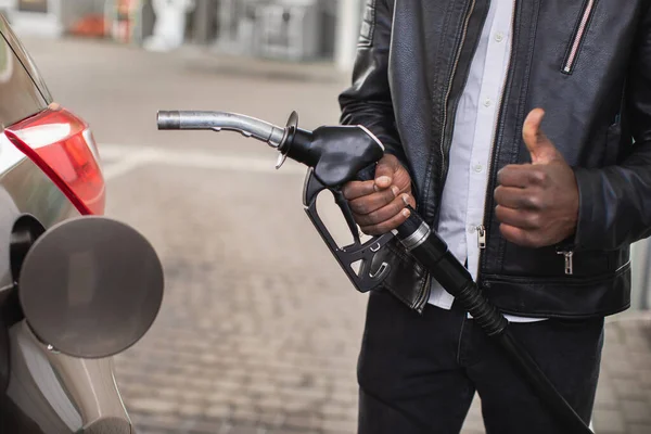 Primo piano immagine ritagliata di uomo dalla pelle nera irriconoscibile in abito casual nero, in piedi all'aperto al distributore di benzina e tenendo in mano l'ugello del fucile a benzina e mostrando il pollice in alto — Foto Stock