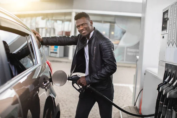 Genç Afro-Amerikalı işadamı benzin istasyonunda modern lüks arabasına yakıt dolduruyor, kameraya gülümsüyor. — Stok fotoğraf