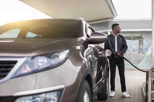 Giovane bel ragazzo barbuto africano in elegante abbigliamento casual che rifornisce la sua auto di lusso e distogliendo lo sguardo mentre in piedi sul distributore di benzina self-service — Foto Stock